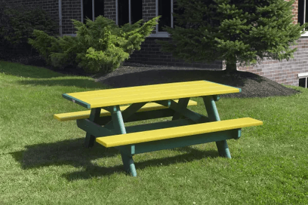 Green and yellow rectangular picnic table on a grassy lawn, near a brick building and trees.